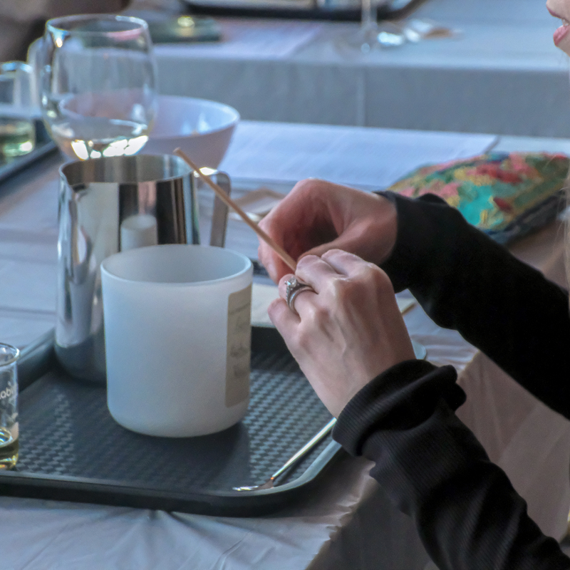 Image of a candle-making class with a cozy atmosphere. Participants are guided by Gloria to create coconut soy wax candles with wooden wicks and non-toxic scents. Includes materials, charcuterie, and a drink of choice (wine, prosecco, or beer). Option to add a second candle or room spray to your experience.